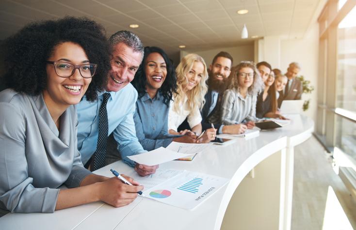 Diverse group of people in business attire leaning over a long desk next to a window smiling