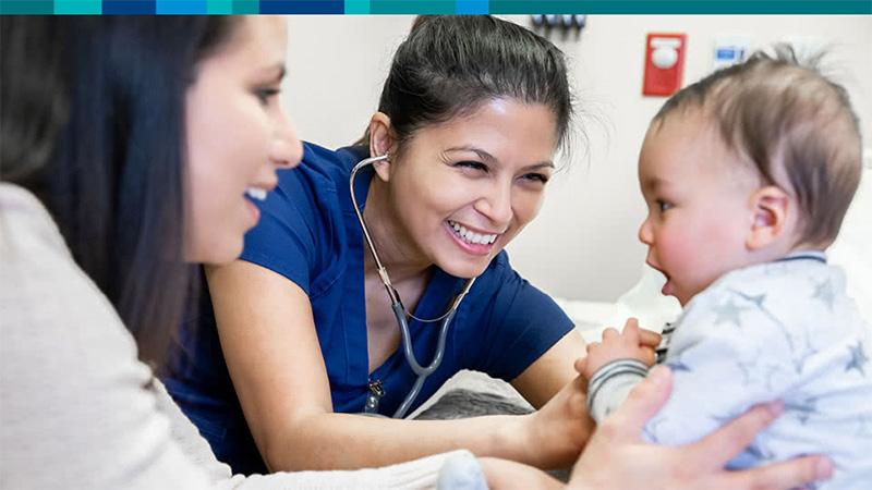 Mother holding young baby in a jumpsuit while a doctor wearing a stethoscope  smiles