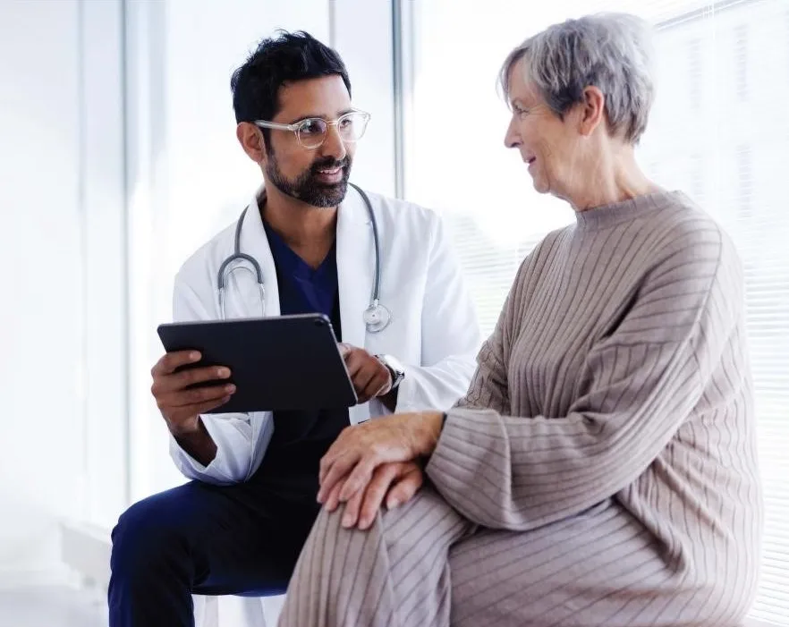 Image of a GP in a white coat sitting with elderly client discussing matters.