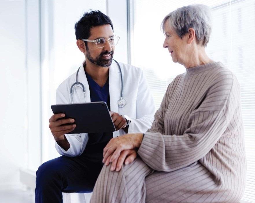 Image of a GP in a white coat sitting with elderly client discussing matters.