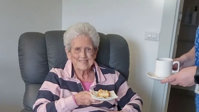 Wendy sitting in an armchair holding a scone being served a cup of tea