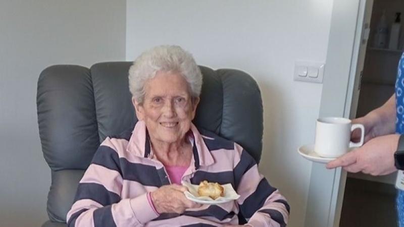 Wendy sitting in an armchair holding a scone being served a cup of tea
