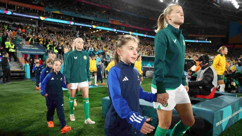 2 Matildas players walking onto a field holding hands of 2 young girls