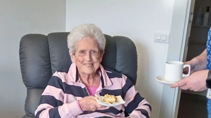 Wendy sitting in an armchair holding a scone being served a cup of tea