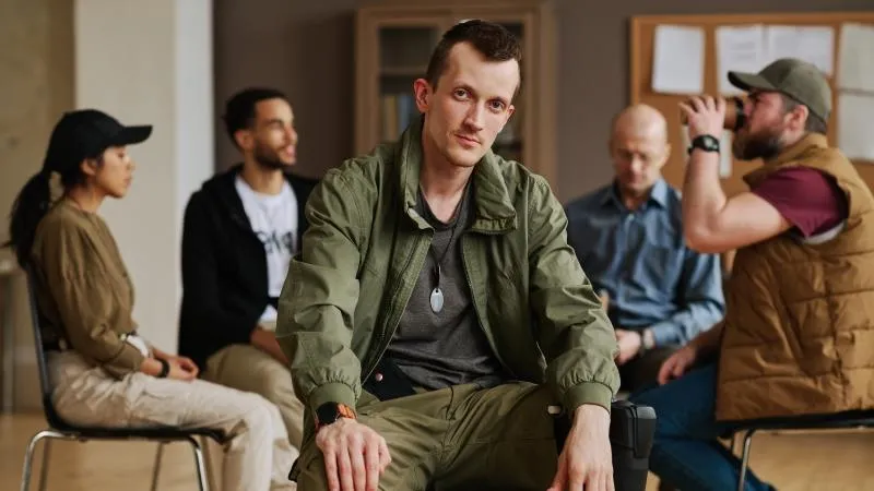 Man in a bomber jacket sitting in front of other people in a support group circle