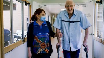 A health care worker has her arm around the arm of a man supporting him while walking. They are in a health care facility. The man is using a walking frame.