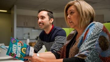 A side on image of two people sitting at a table. One is holding the ‘a journey into sorry business’ booklet.