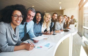 Diverse group of people in business attire leaning over a long desk next to a window smiling