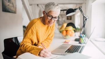 Woman wearing a sweater and glasses sitting at a desk with a laptop in a bright room