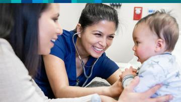 Mother holding young baby in a jumpsuit while a doctor wearing a stethoscope  smiles