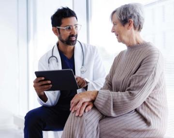 Image of a GP in a white coat sitting with elderly client discussing matters.