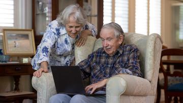 Elderly man in an armchair looking at a laptop with an elderly woman looking over his shoulder smiling
