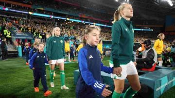 2 Matildas players walking onto a field holding hands of 2 young girls