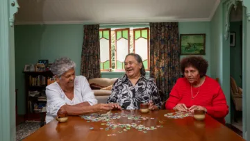 Three elderly women sitting around a table doing a puzzle and chatting
