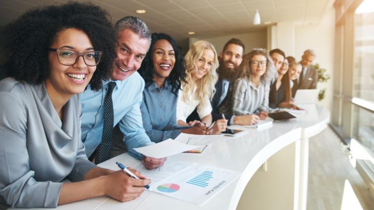 Diverse group of people in business attire leaning over a long desk next to a window smiling