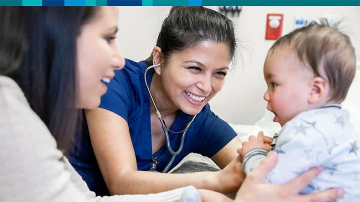 Mother holding young baby in a jumpsuit while a doctor wearing a stethoscope  smiles
