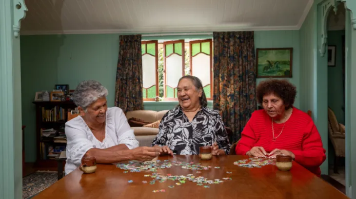 Three elderly women sitting around a table doing a puzzle and chatting