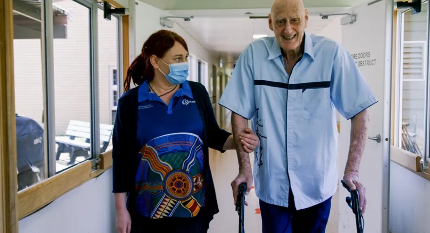 A health care worker has her arm around the arm of a man supporting him while walking. They are in a health care facility. The man is using a walking frame. 