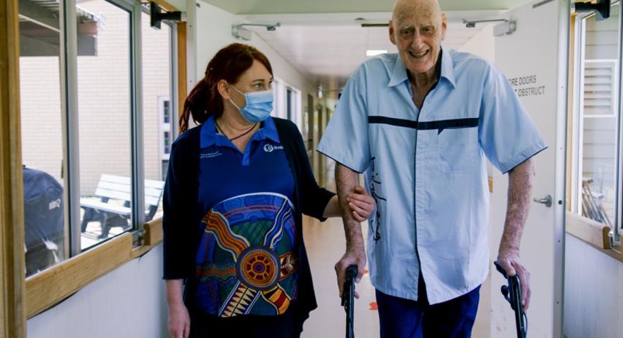 A health care worker has her arm around the arm of a man supporting him while walking. They are in a health care facility. The man is using a walking frame. 