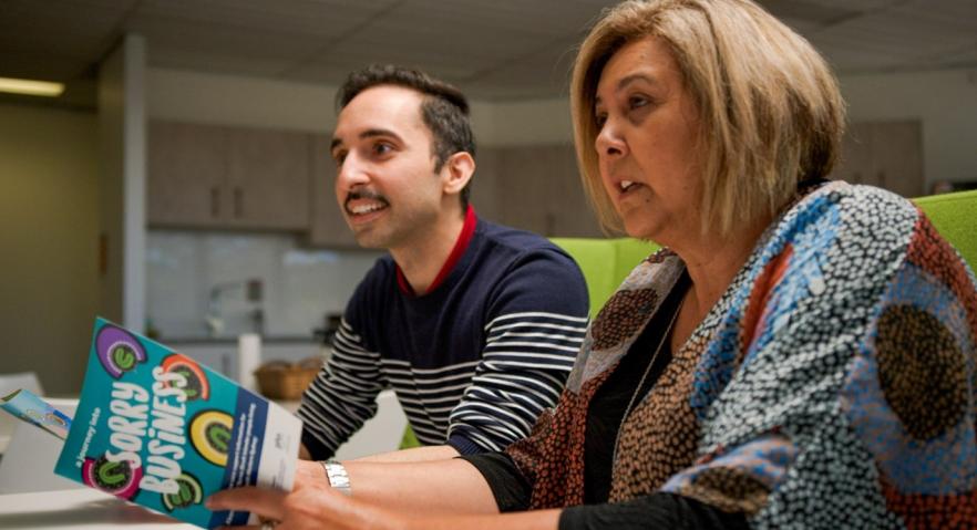 A side on image of two people sitting at a table. One is holding the ‘a journey into sorry business’ booklet. 